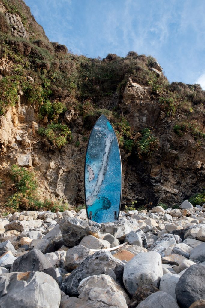 Marte 01, tabla de surf de SONEK, de pie entre los cantos rodados, al pie de un acantilado