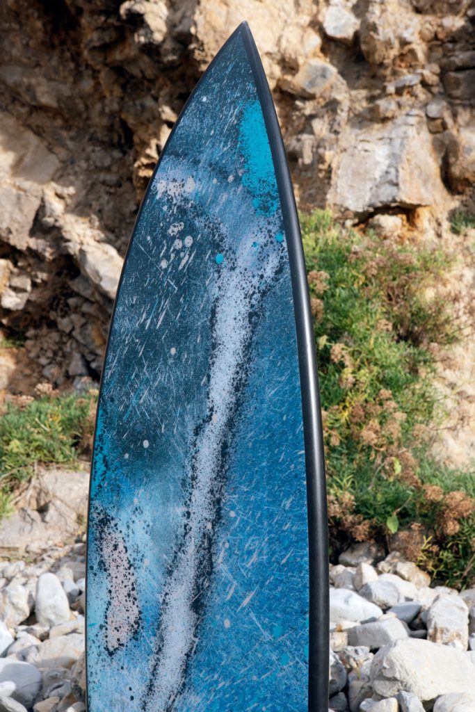 Marte 01, tabla de surf de SONEK, de pie entre las piedras y la vegetación del acantilado
