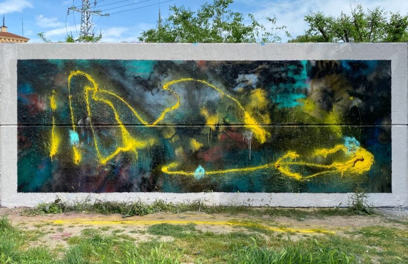 Mural abstracto de SONEK en la Abstract Session: fondo en azules, turquesas y negros con una línea amarilla de spray, en un panel enmarcado en blanco sobre un muro de hormigón, con césped delante, una torre eléctrica y árboles detrás.