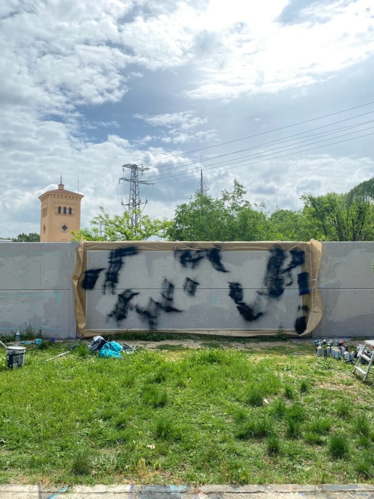 El panel enmarcado con cinta de carrocero y las primeras manchas negras de spray sobre fondo claro, con una torre de ladrillo y árboles detrás.