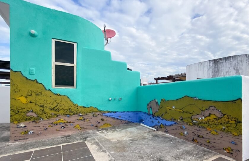 Mural de paisaje de SONEK: colinas verdes y ocres, rocas y un mar azul con un arco de roca recorren la base de dos muros turquesa, con una ventana y un cielo nublado detrás.