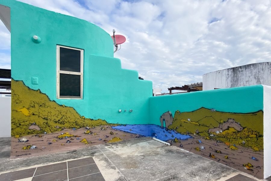 Mural de paisaje de SONEK: colinas verdes y ocres, rocas y un mar azul con un arco de roca recorren la base de dos muros turquesa, con una ventana y un cielo nublado detrás.