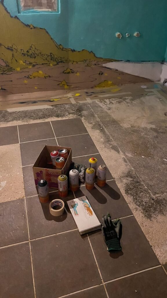 De noche, una caja de sprays, cinta y guantes en el suelo, con el paisaje ya empezado en la pared.