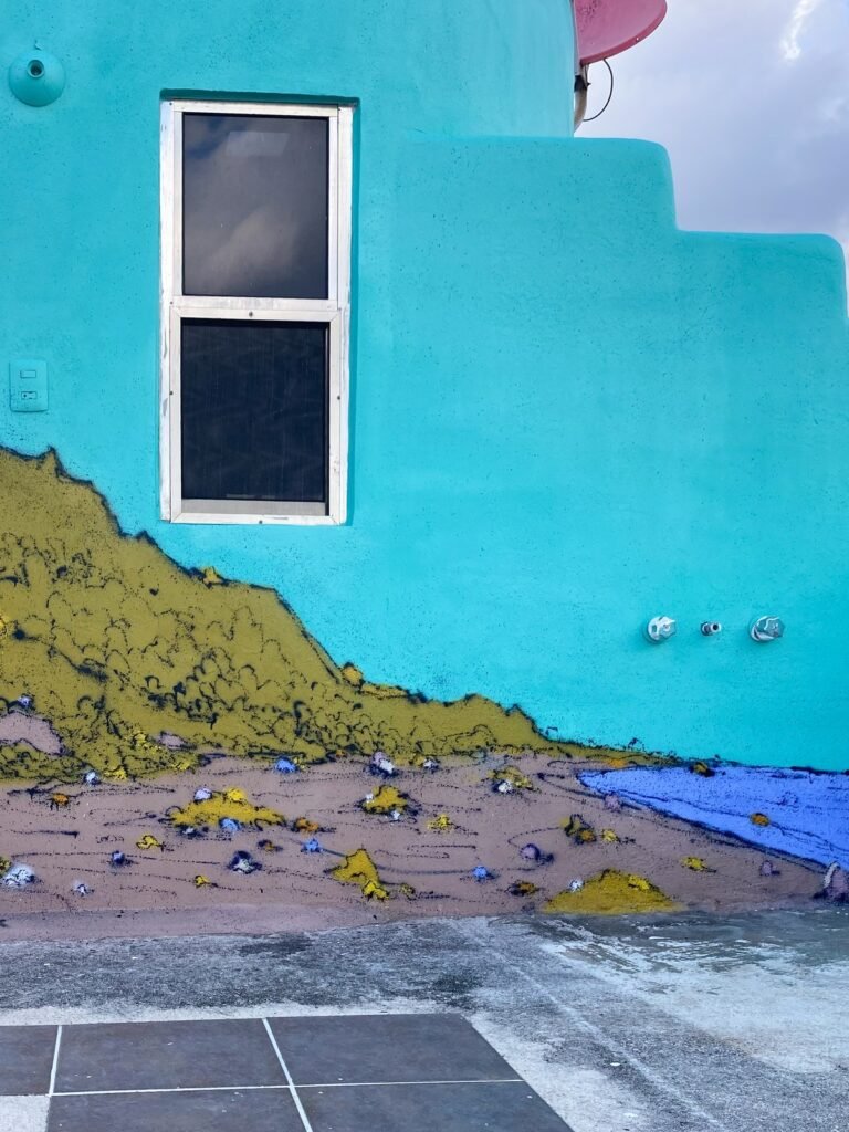 Una ladera verde baja desde debajo de una ventana hasta el mar azul, sobre la pared turquesa.