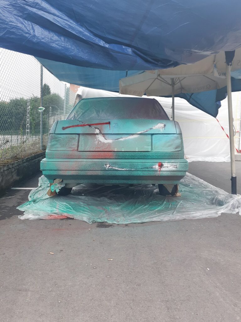 Trasera del coche bajo una lona azul, con los primeros trazos blancos y rojos sobre el verde