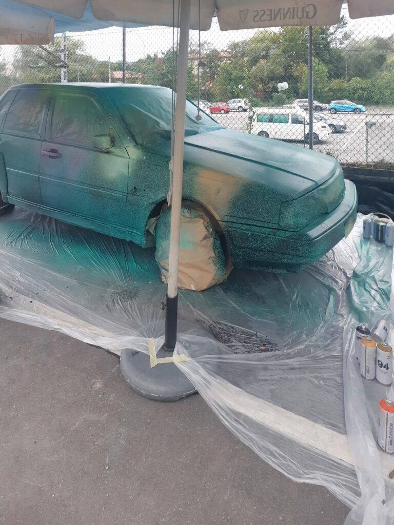 Coche tapado con plásticos bajo una sombrilla, con la primera capa de spray verde y los botes al lado