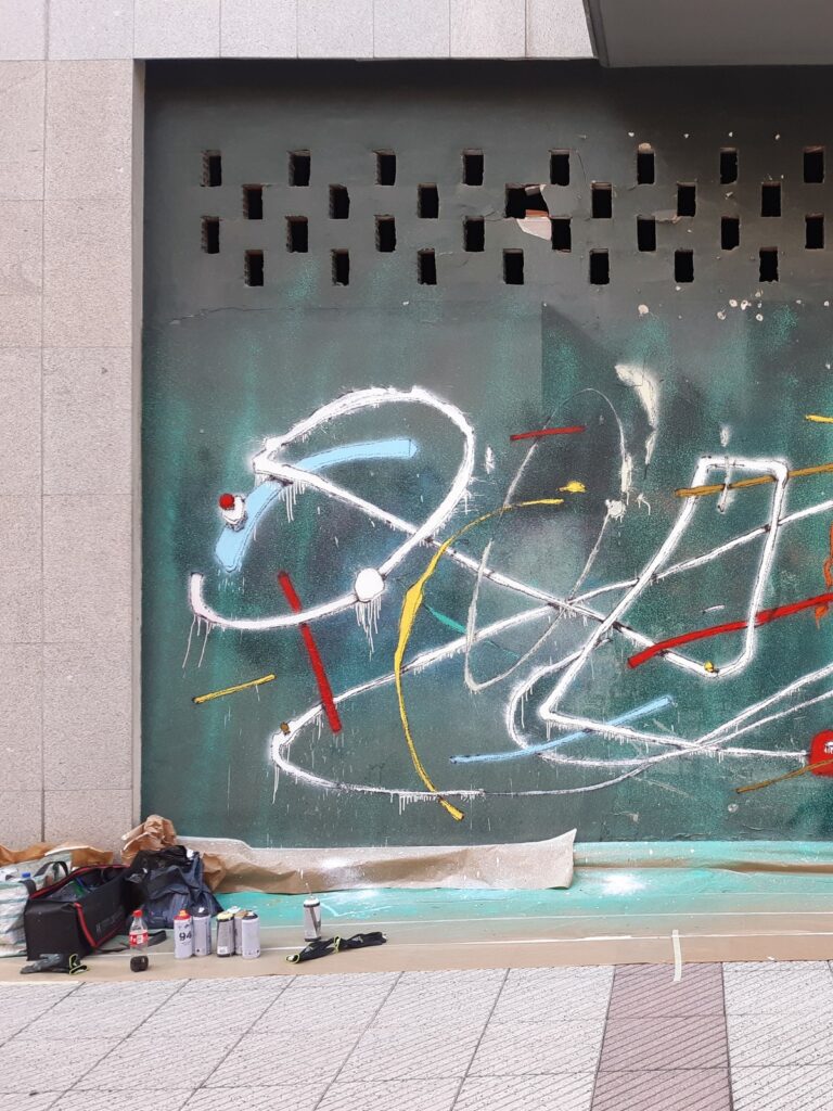 Persiana de un local pintada con trazos blancos, azules, amarillos y rojos sobre verde, con los botes de spray y las bolsas en la acera