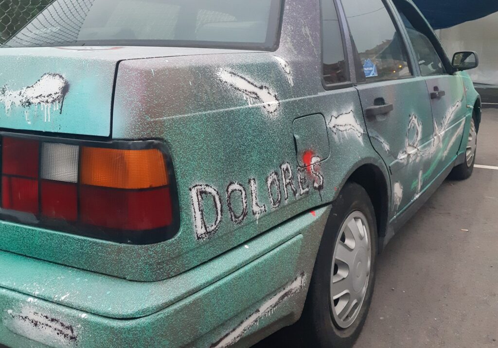 Trasera de un coche pintado de verde con salpicaduras, trazos blancos y rojos y el nombre Dolores escrito en blanco en el lateral