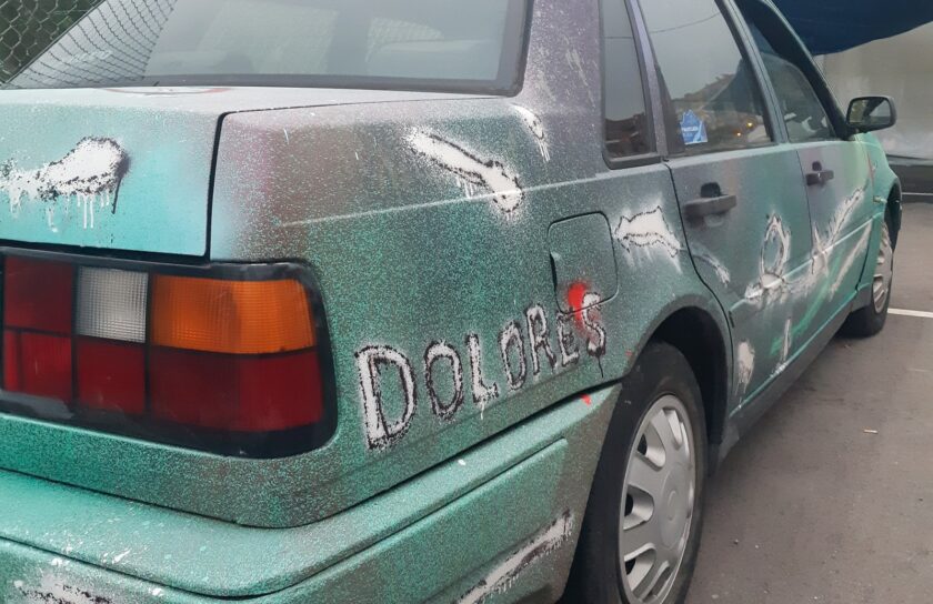 Trasera de un coche pintado de verde con salpicaduras, trazos blancos y rojos y el nombre Dolores escrito en blanco en el lateral