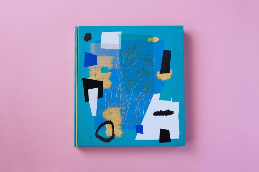 Cubierta turquesa del libro El Mundo del Hogar intervenida con recortes de papel negro, azul y blanco y trazos de cera, sobre fondo rosa