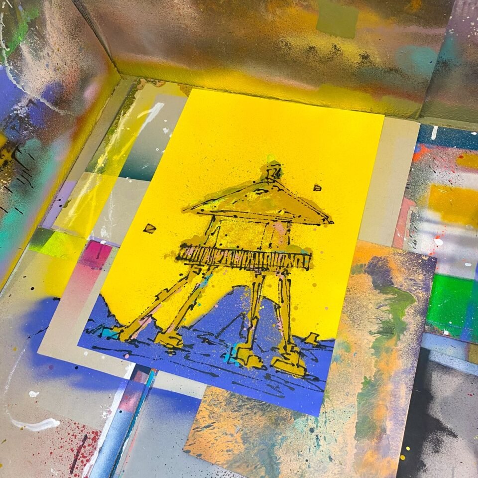 Un hórreo en negro sobre papel amarillo, en el suelo del taller entre papeles de colores