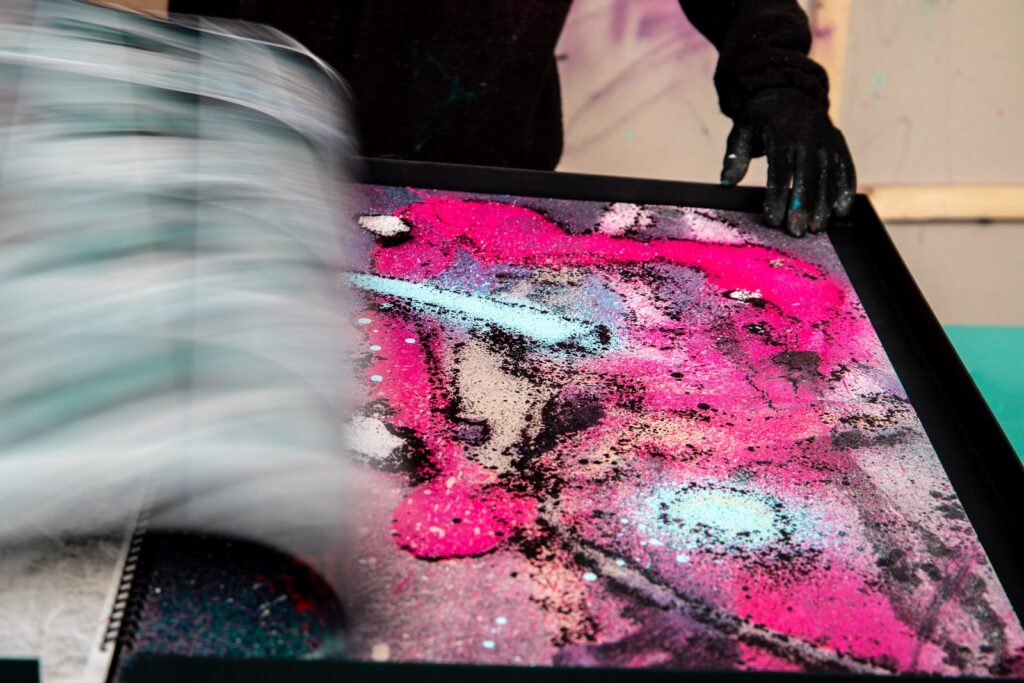 Hojas del catálogo pasando en movimiento sobre una página intervenida a spray en rosa, con las manos a los lados del libro