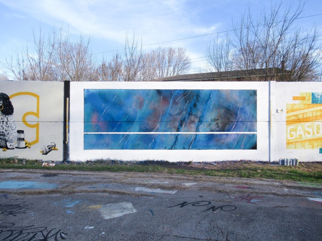 El mural azul enmarcado en blanco, entre piezas de otros artistas en un muro libre, con el suelo pintado delante