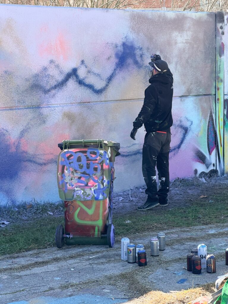 SONEK pintando con spray el fondo lila y rosa del mural, con el primer trazo azul, junto a un contenedor lleno de pintadas y botes de spray en el suelo