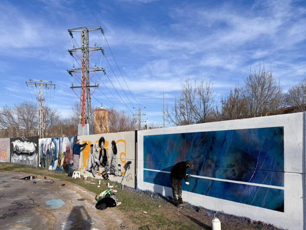 El muro libre de lejos, con piezas de otros artistas a la izquierda y SONEK pintando la línea blanca del mural azul, bajo torres de alta tensión