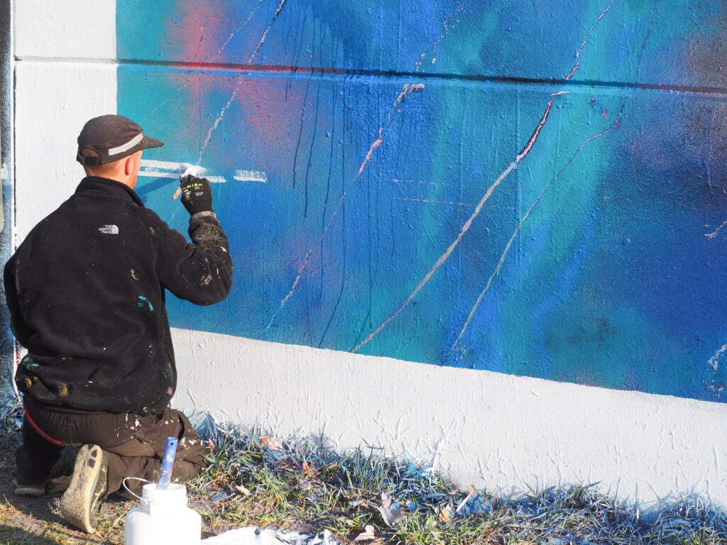 SONEK, de rodillas y de espaldas, pintando con pincel la línea blanca sobre el azul del mural