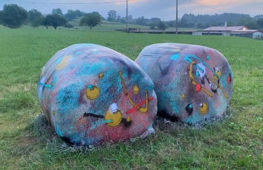 Las dos bolas de silo terminadas, pintadas en turquesa, rosa, negro, amarillo y blanco, en un prao de Porrúa bajo un cielo naranja y rosado al atardecer.