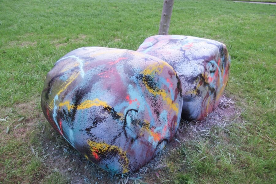 Dos bolas de silo pintadas en turquesa, amarillo, negro y blanco sobre la hierba, con un tronco delgado detrás.