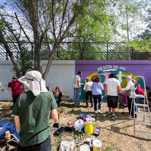 Plano general del taller en Soto del Real: jóvenes y mayores pintan el mural One Earth One Love, con el material en el suelo.