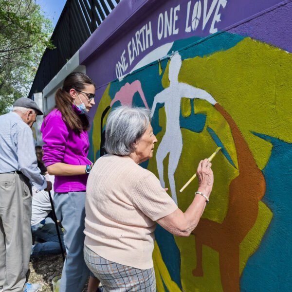 Una mujer mayor pinta con pincel una figura del mural One Earth One Love, junto a jóvenes y otro participante mayor.