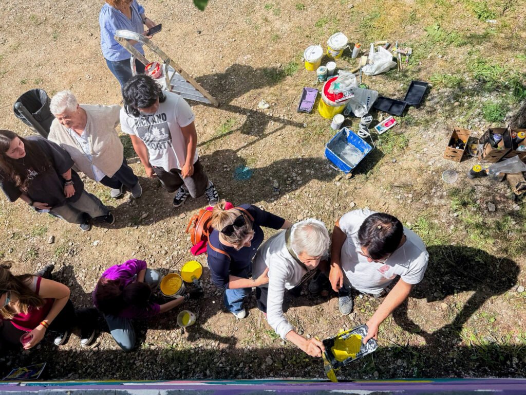 Jóvenes y mayores pintan tableros en el suelo, vistos desde arriba, entre botes y bandejas de pintura.