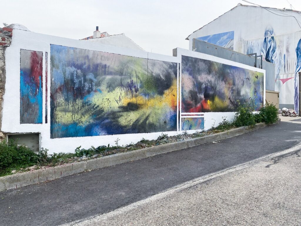 Mural terminado visto en diagonal desde la calle, con manchas azules, amarillas, rosas y rojas sobre un muro blanco