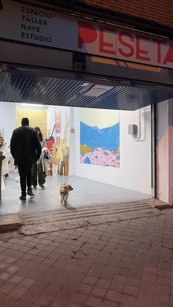 La entrada de Espacio Peseta de noche: el letrero de Peseta arriba, gente y un perro entrando y el mural al fondo.