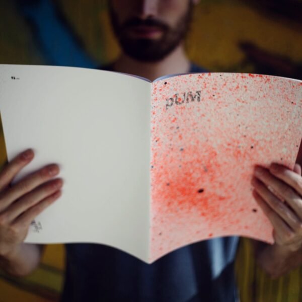 Una persona sostiene abierto el fanzine Pum, con la portada salpicada de spray, delante de un mural.