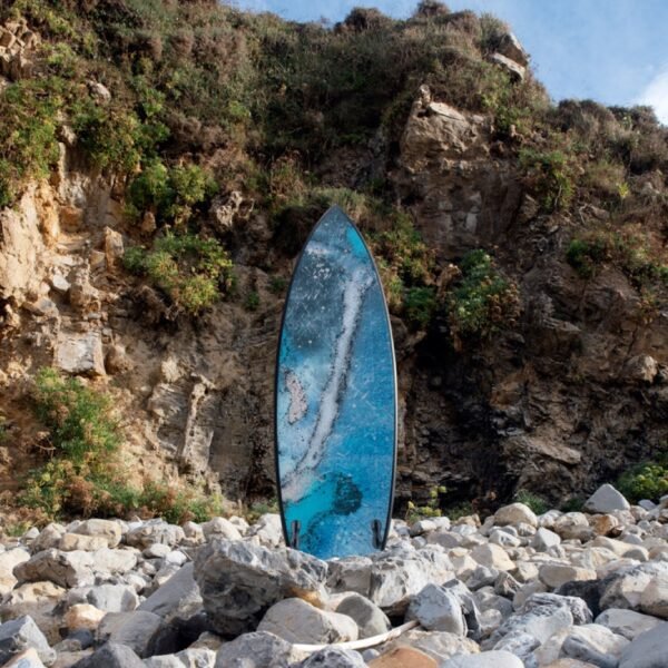 Tabla de surf pintada en azules por Sonek, apoyada en una playa de cantos rodados