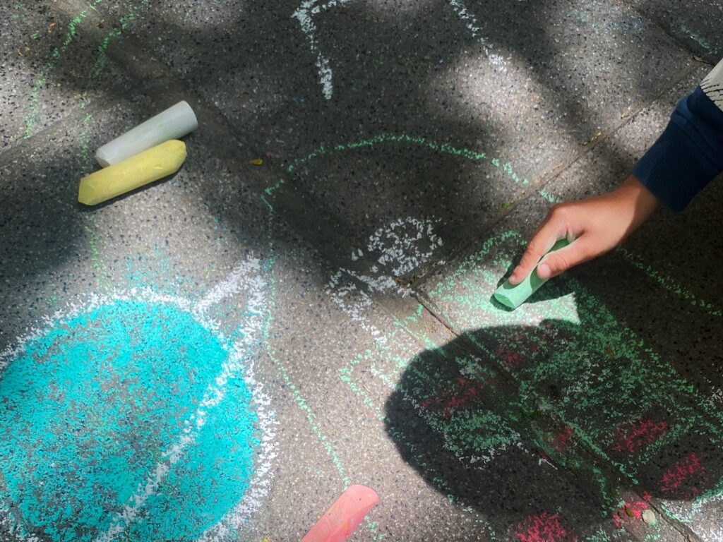Vista cenital de una mano rellenando con una tiza verde, con barras de tiza sueltas y un círculo turquesa dibujado en el suelo