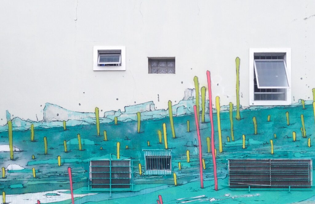 Mural Brotes: palos altos amarillos y rosas sobre un mar turquesa en la parte baja de una fachada blanca, con tres ventanas y rejillas.