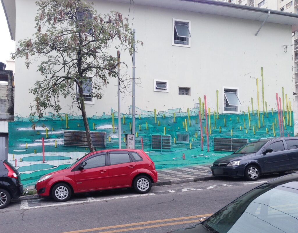 La fachada terminada vista desde la calle: palos amarillos y rosas sobre el turquesa, con un árbol y un coche rojo delante.