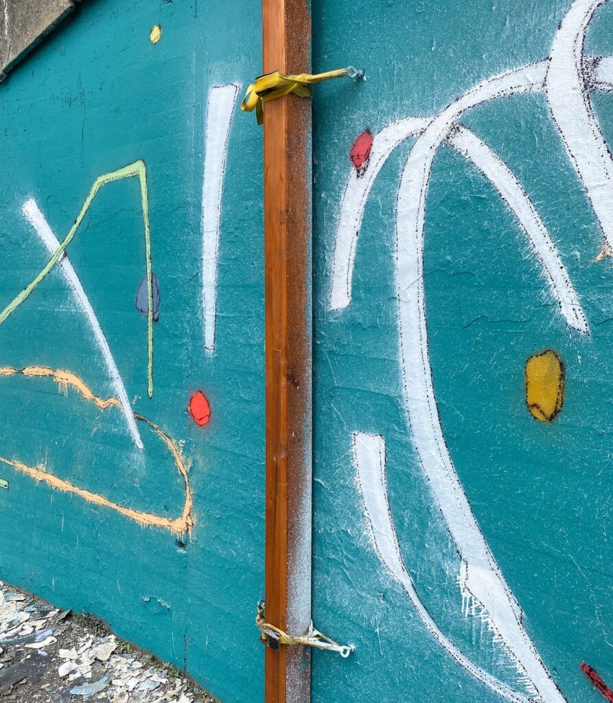 Detalle del muro con trazos blancos, un óvalo amarillo y un poste de madera atado con una cinta amarilla.