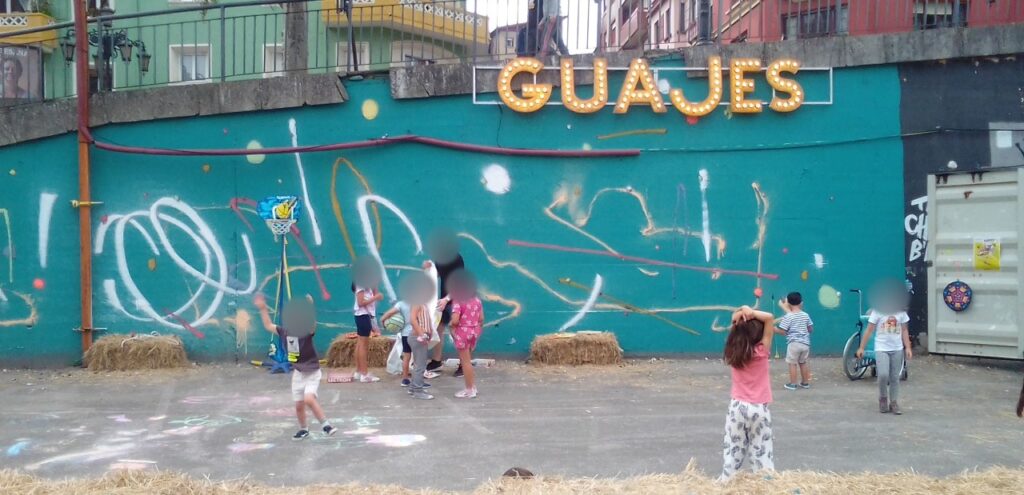 Niños jugando con un aro de baloncesto y pañuelos delante del muro turquesa, bajo el rótulo «GUAJES». Las caras están difuminadas.