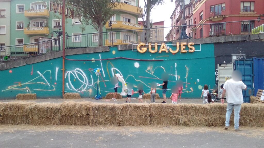 El muro turquesa con el rótulo «GUAJES» encima, pacas de paja delante y niños jugando, en la zona de guajes del Kuivi Pop Up. Las caras están difuminadas.