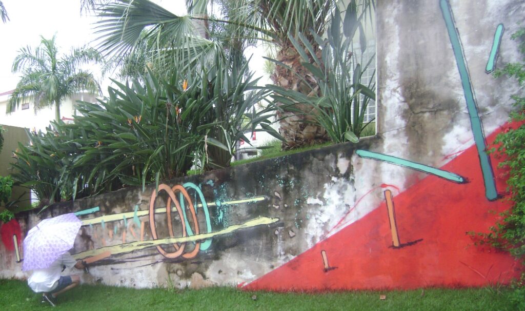 Una persona agachada, con paraguas, pinta en la parte baja del muro; ya están los aros, las barras amarillas y el triángulo rojo, con palmeras y plantas detrás.
