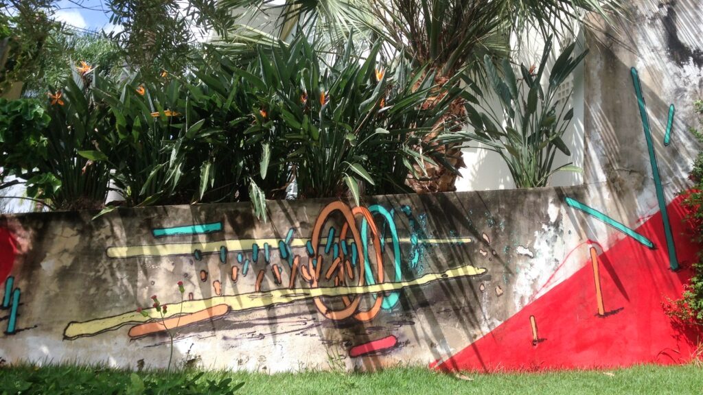 El muro con los aros naranjas y turquesas, las barras amarillas y el triángulo rojo, con sol, sombras de palmera y flores naranjas delante.