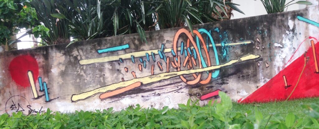 El muro visto de frente, con los aros naranjas y turquesas, las barras amarillas, un círculo rojo a la izquierda y el triángulo rojo a la derecha, tras unas plantas verdes.