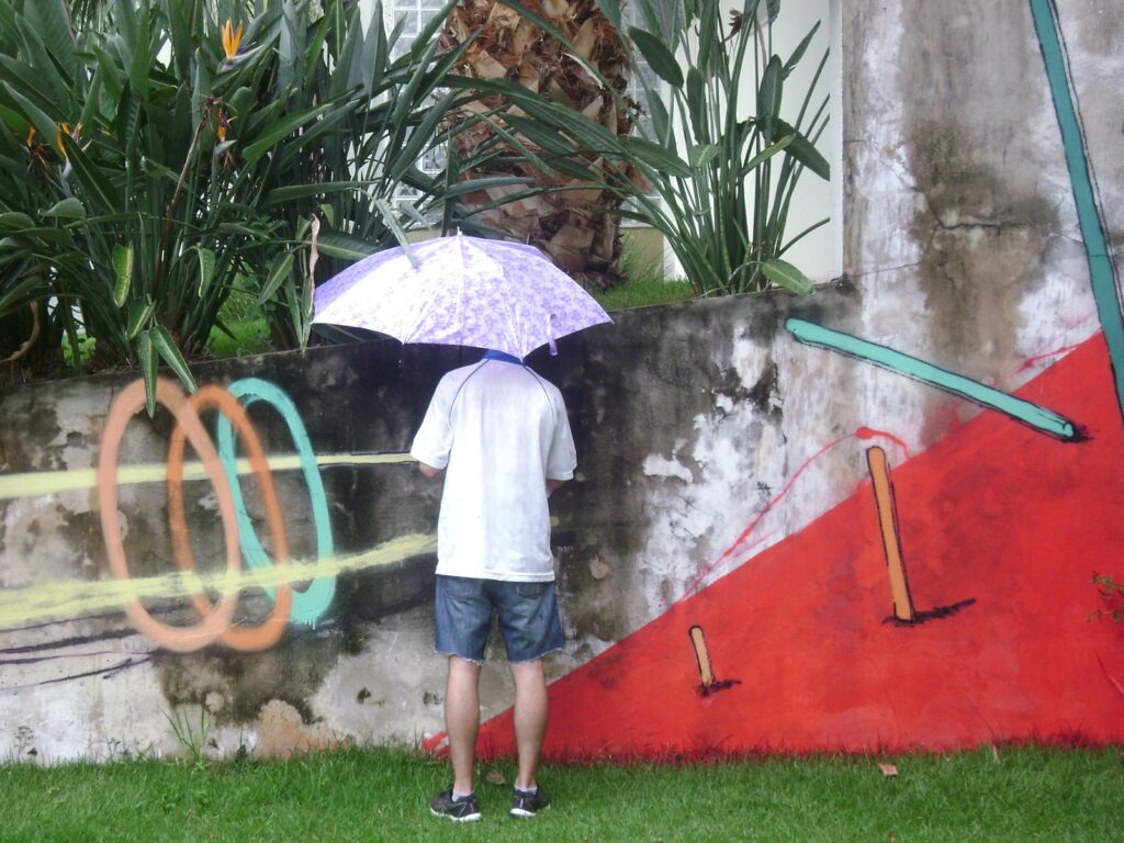 Una persona de espaldas, con paraguas, pinta sobre un muro bajo de un jardín: a la izquierda unos aros y a la derecha un triángulo rojo.