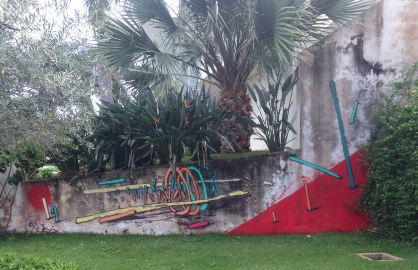 Mural Coche: aros naranjas y turquesas sobre barras amarillas, un círculo rojo y un triángulo rojo en un muro bajo de jardín, con una palmera y un olivo detrás.