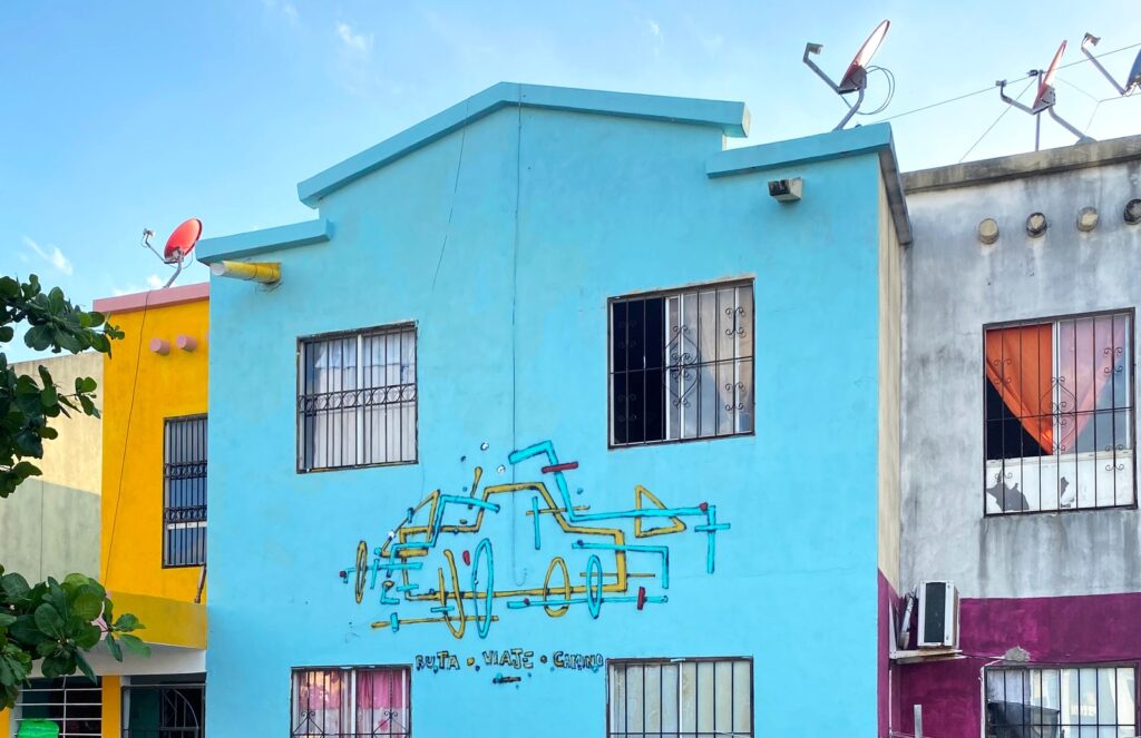 Mural En ruta: un coche dibujado con líneas amarillas y turquesas y el texto «Ruta · Viaje · Camino» sobre la fachada azul de una casa de dos plantas.