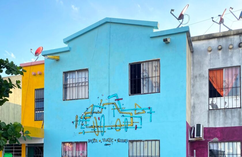 Mural En ruta: un coche dibujado con líneas amarillas y turquesas y el texto «Ruta · Viaje · Camino» sobre la fachada azul de una casa de dos plantas.
