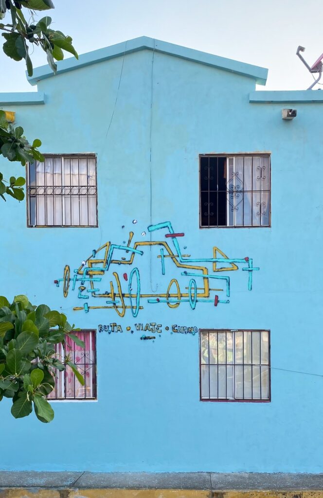 Fachada azul de dos plantas con el coche en amarillo y turquesa y el texto «Ruta · Viaje · Camino» entre las ventanas.