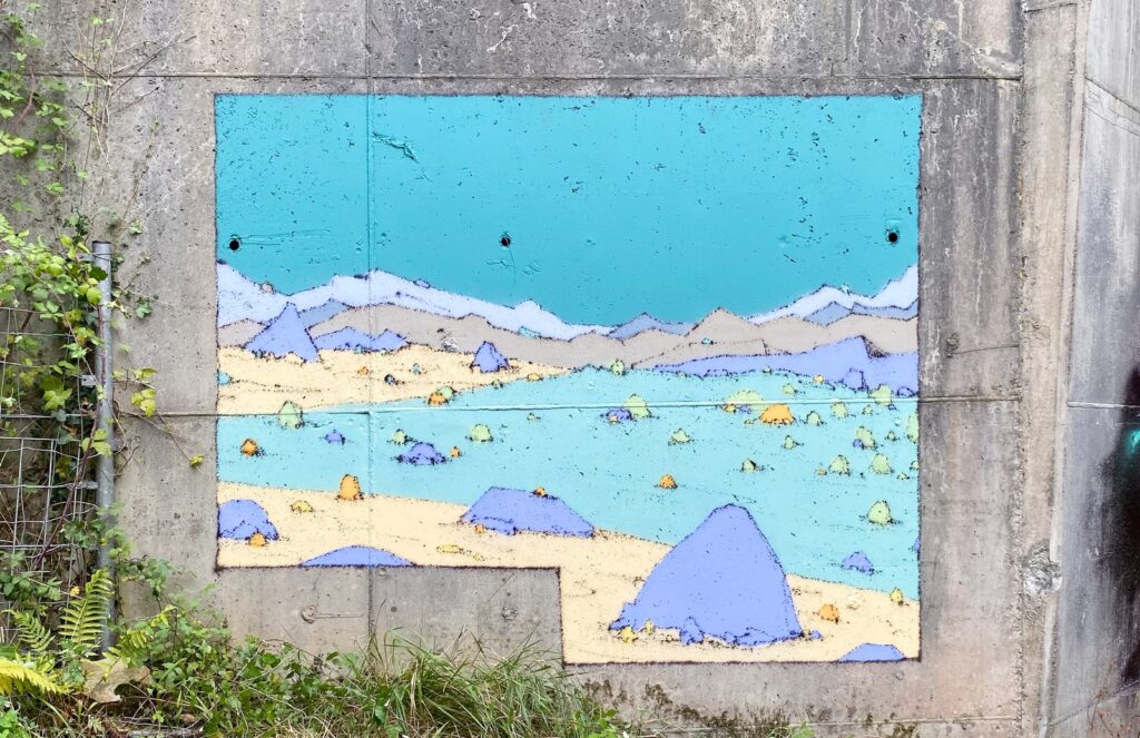 Mural Entrepraos: paisaje de montañas azules y arena pintado sobre un muro de hormigón, entre la hierba.