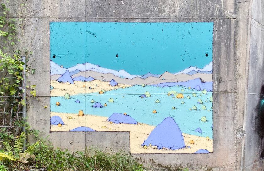 Mural Entrepraos: paisaje de montañas azules y arena pintado sobre un muro de hormigón, entre la hierba.