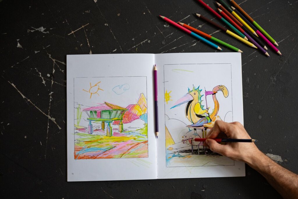 El cuadernillo abierto por un hórreo y un pájaro, con una mano coloreando y lápices de colores alrededor, sobre una mesa negra