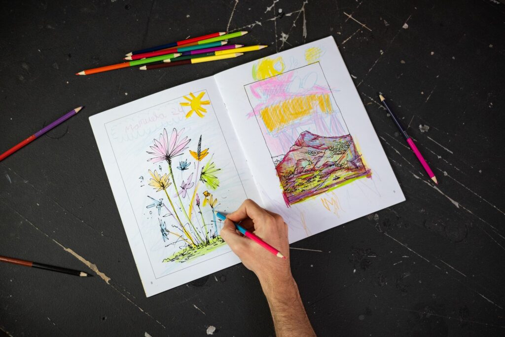 El cuadernillo abierto por unas flores y una montaña, con una mano coloreando, sobre una mesa negra