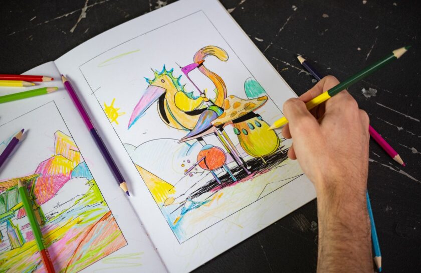 Una mano colorea con un lápiz amarillo un pájaro del cuadernillo, sobre una mesa negra con lápices de colores