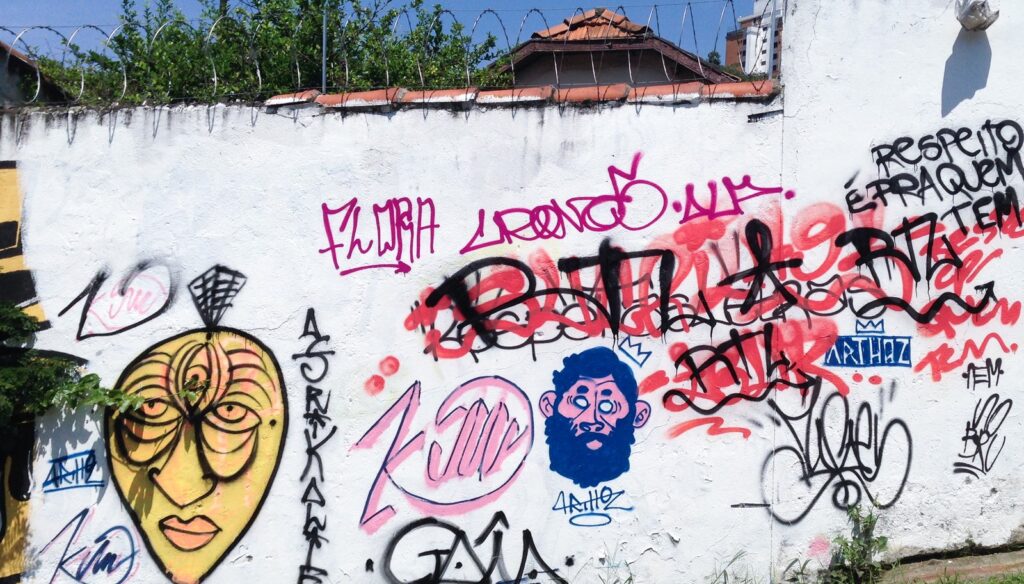 La pared blanca antes de pintar, con otros grafitis, entre ellos una máscara amarilla a la izquierda, bajo un cielo azul y alambre de espino.