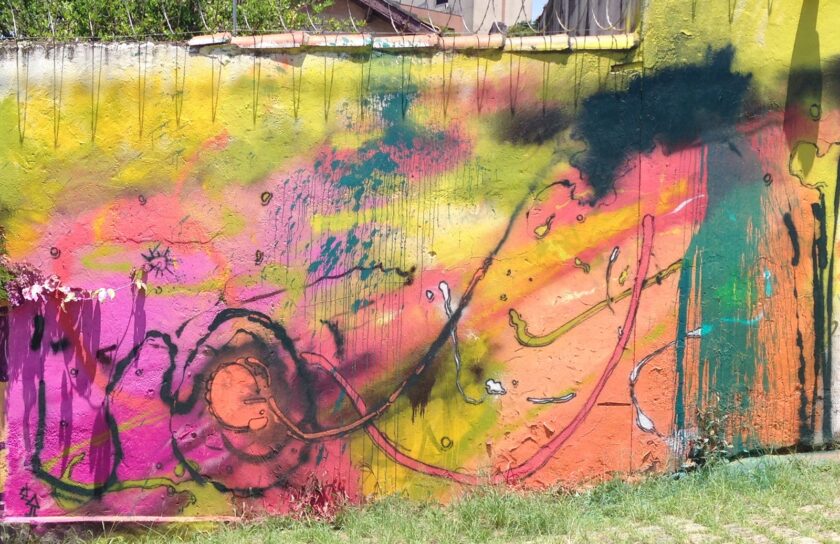 Mural Saudade: manchas rosas, amarillas, naranjas y verdes con trazos oscuros y una espiral en el centro, sobre una pared larga con alambre de espino por arriba y hierba al pie.